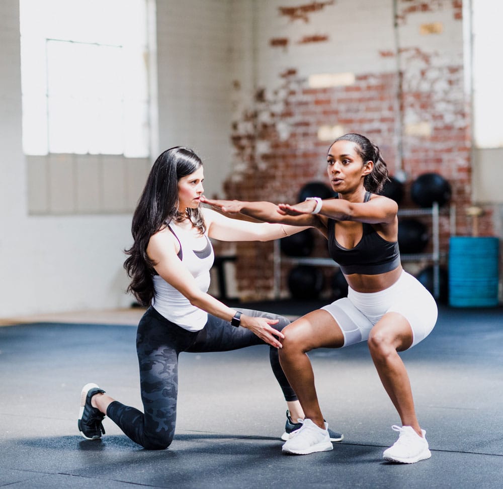 Coach sportive brune guide une femme réalisant un squat, bras tendus, pour corriger la posture en salle de gym.