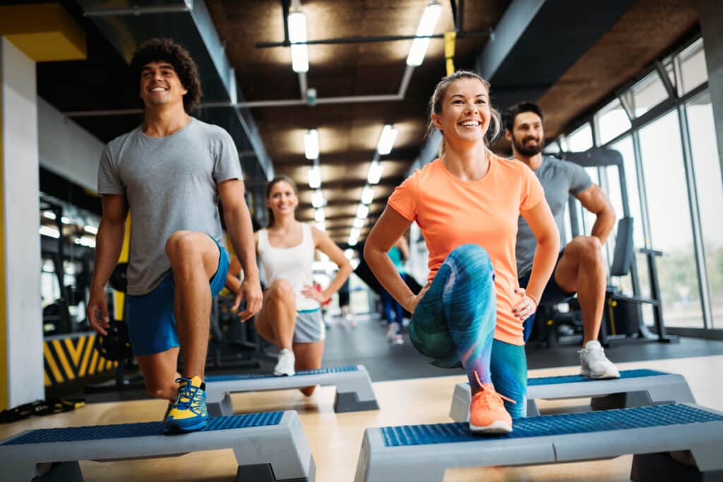 Cours de Step: Entraînement en Groupe Joyeux au Gym Quatre personnes souriantes font des step-ups sur des plateformes aérobic dans une salle de sport lumineuse.