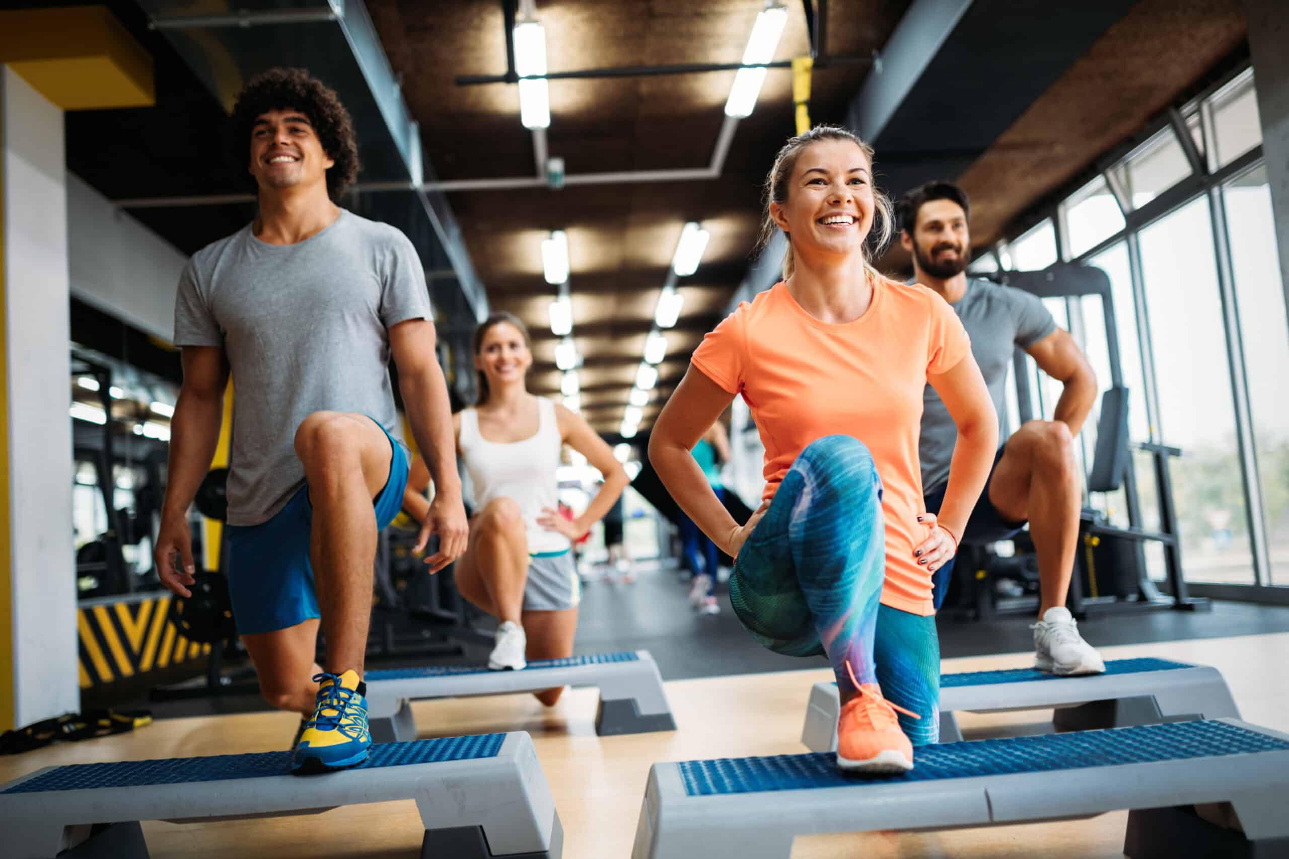 Quatre personnes souriantes font des step-ups sur des plateformes aérobic dans une salle de sport lumineuse.