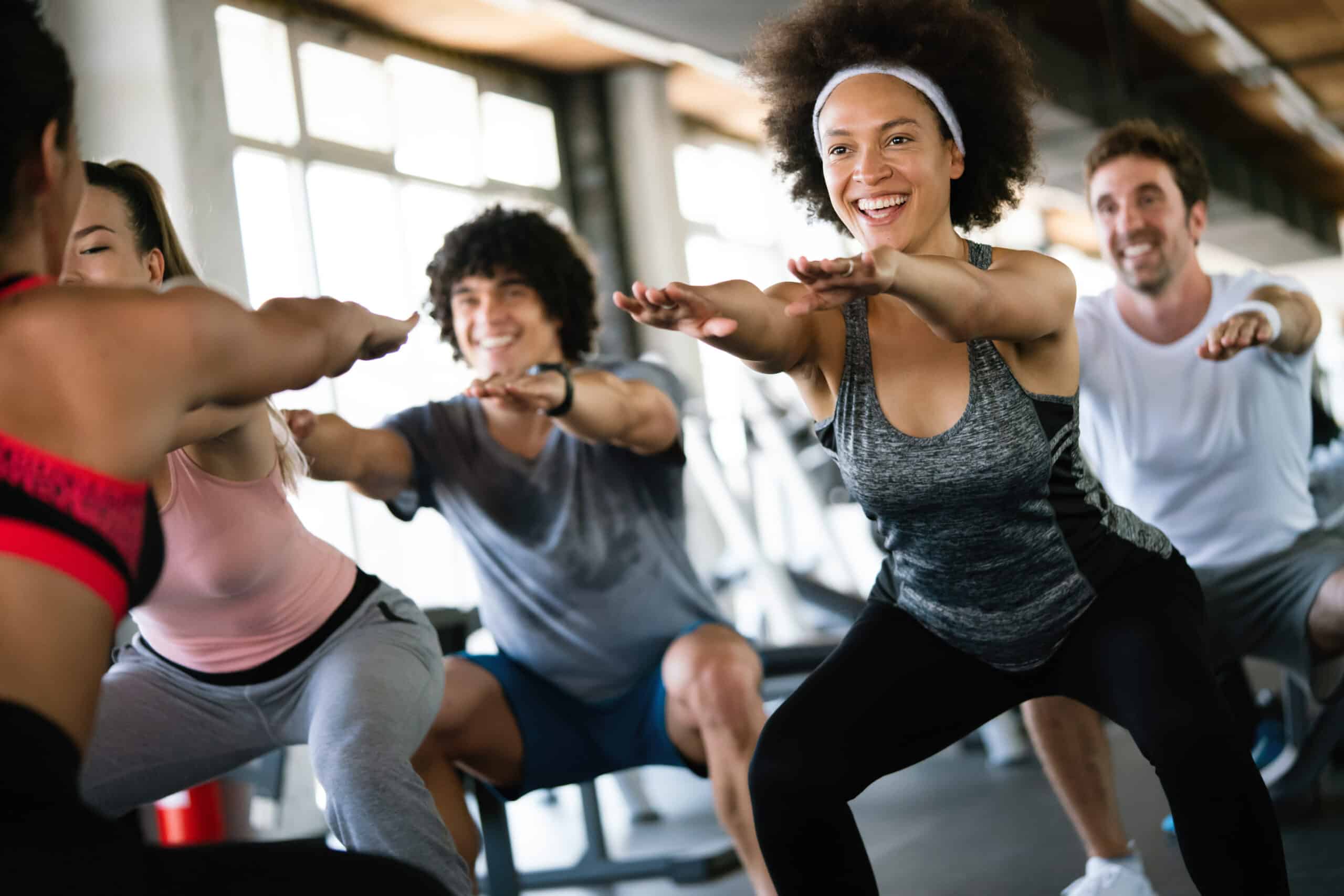 Groupe souriant effectuant des squats en gym. Une femme joyeuse aux cheveux afro au premier plan. Entraînement sportif en groupe.