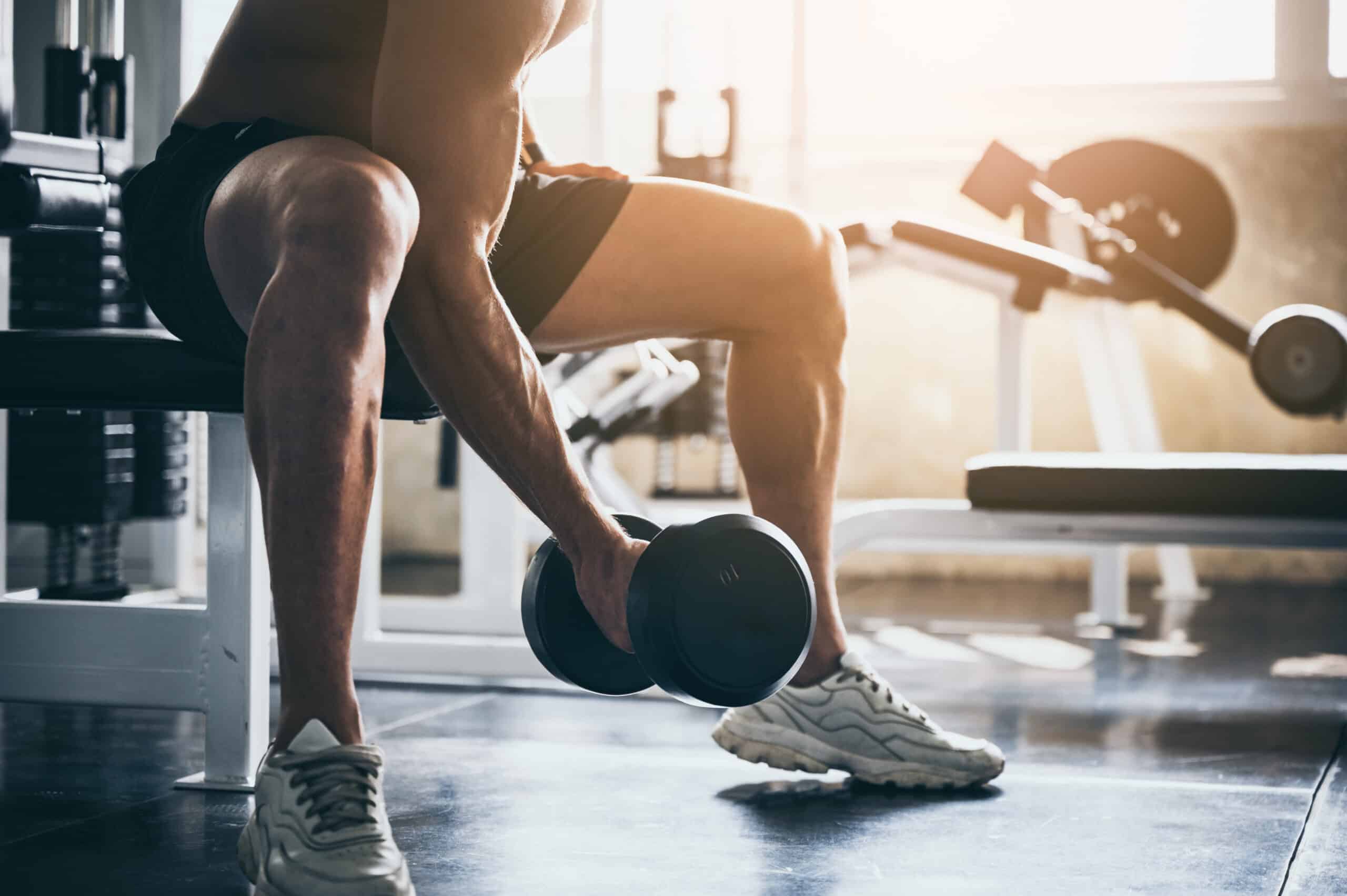 Homme musclé concentré soulevant un haltère dans une salle de sport lumineuse, jambes et bras tendus. Entraînement de force intense.