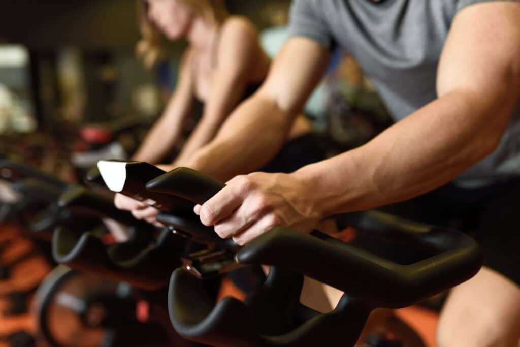 Homme et femme en plein entraînement de vélo spinning en salle de sport. Les mains musclées de l'homme agrippent le guidon.