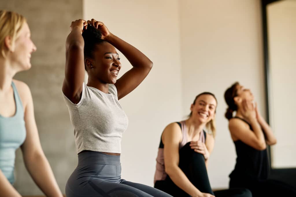 Femme noire souriante ajustant ses cheveux en chignon. Cours de sport avec amies, ambiance joyeuse et bien-être.