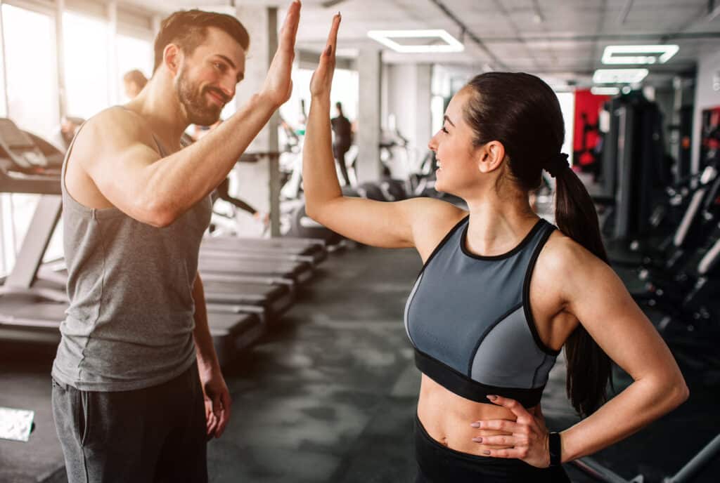 Homme et femme souriants se donnant un high-five dans une salle de sport moderne. Scène de succès et motivation après l'entraînement.