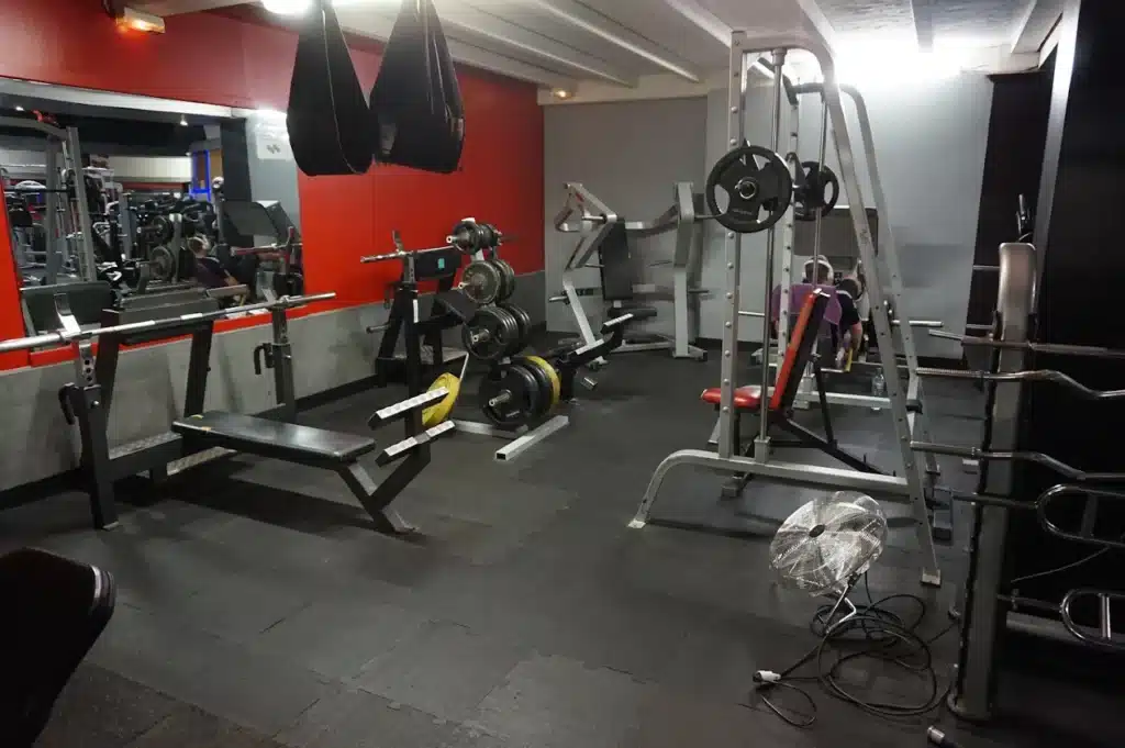 Salle de sport équipée: banc de musculation, Smith machine, racks de poids, et ventilateur. Un utilisateur se reflète dans le miroir. Murs rouges et gris.
